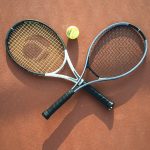 Two tennis rackets and a ball arranged on a clay court, casting shadows.