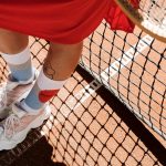 Close-up view of tennis player’s feet on a clay court, highlighting white sneakers and colorful socks.
