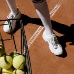 Close-up of tennis shoes and ball basket on a clay court, ready for play.
