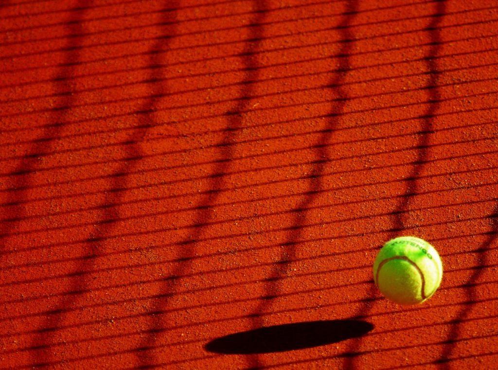 A vibrant tennis ball casting a shadow on a sunlit clay court with net shadows.
