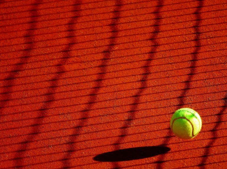 A vibrant tennis ball casting a shadow on a sunlit clay court with net shadows.